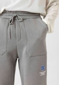 Grijze joggingbroek met een elastische tailleband en trekkoord. Beschikt over twee zijzakken, een blauwe graphic op de linkerdij en een gestructureerde stof.
