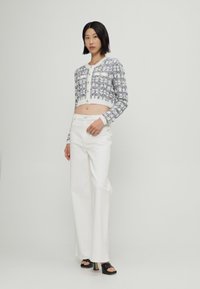 Cardigan court au style crochet dans des couleurs pastel avec des boutons dorés, associé à un pantalon blanc à jambes larges et des sandales noires à talons bloc.