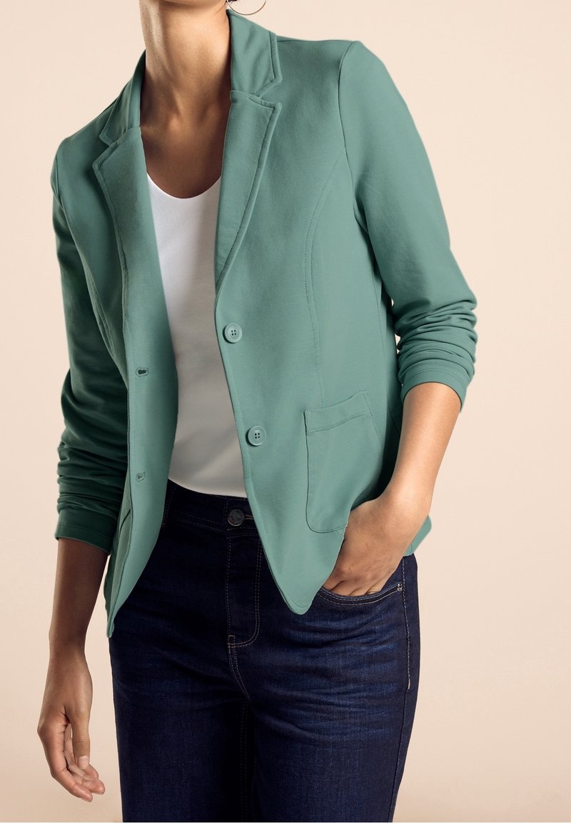 Personne portant un blazer vert clair sur une chemise blanche et un jean bleu foncé, une main dans la poche, sur un fond beige.