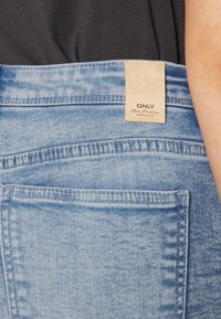 Vaaleansiniset farkut korkealla vyötäröllä, jossa on ommeltu takatasku ja vaaleanruskea etiketti, jossa lukee "ONLY Blue Denim." Kankaan pinta on tekstuurinen.