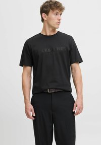 Crna pamučna majica s teksturiranim logotipom "JACK & JONES PREMIUM" sprijeda. Kratki rukavi i okrugli izrez. Uparena s crnim hlačama.