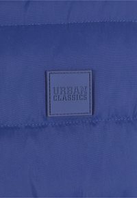 Mėlynas paminkštintas audinys su tekstūruotu paviršiumi. Ant audinio yra kvadratinė juoda etiketė su iškaltu užrašu „URBAN CLASSICS“. Siūlės yra matomos.