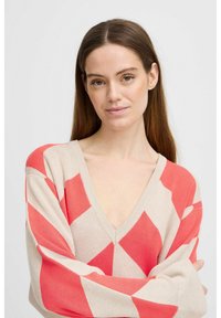 Pull beige avec un motif de losanges corail, présentant un décolleté en V profond, des manches surdimensionnées et une texture en maille douce.
