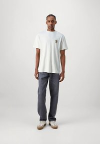 Hellgraues Herren-T-Shirt mit kurzen Ärmeln, rundem Ausschnitt und kleinem gesticktem Logo auf der Brust, kombiniert mit dunkelgrauen Jeans in geradem Schnitt.