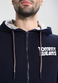 Felpa con zip di colore blu navy con cappuccio foderato in grigio, con logo bianco "TOMMY JEANS" e lacci intrecciati rossi e bianchi. Materiale in misto cotone.