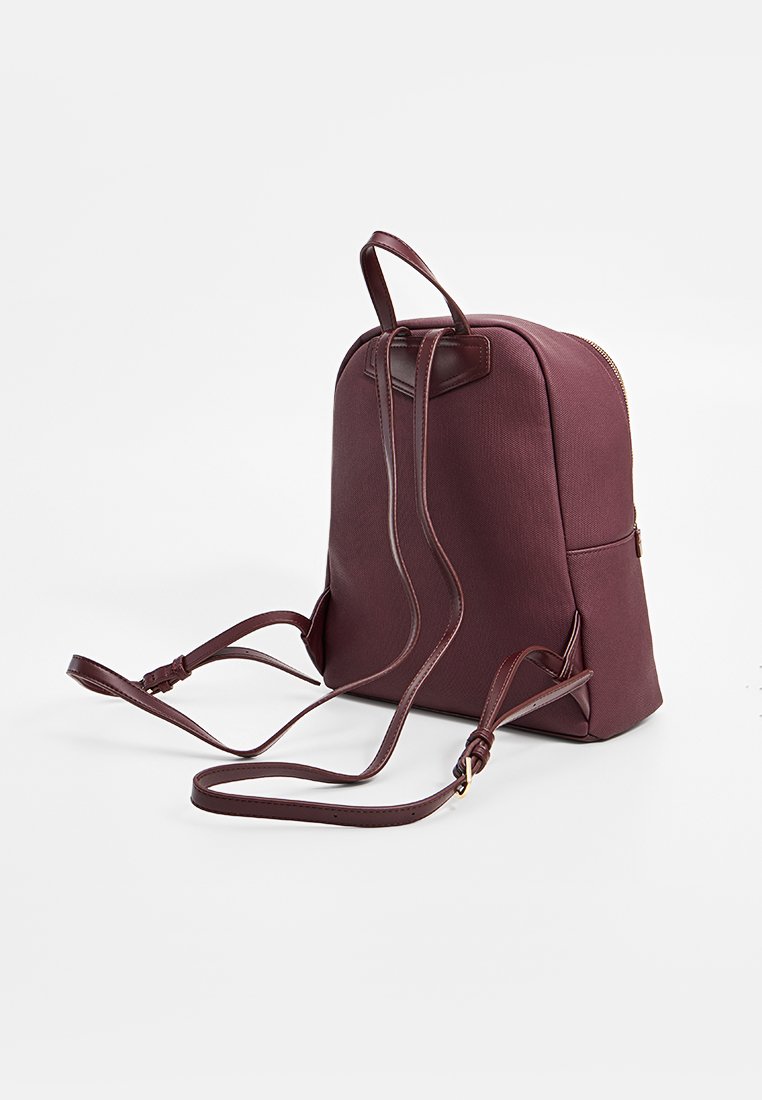 Kompakt burgunderfarget ryggsekk med glatt stoff, justerbare lærstropper og gullglidelås på hvit bakgrunn.