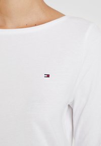 Nahaufnahme einer Person, die ein weißes Langarm-Shirt mit einem kleinen rechteckigen gestickten Logo in Rot, Weiß und Marineblau auf der Brust trägt.