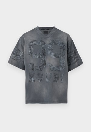 Mężczyzna stoi, ubrany w czarną, zniszczoną, oversize'ową koszulkę z nadrukiem "99", jasne, podarte jeansy z naszywkami oraz czarne trampki na białym tle.