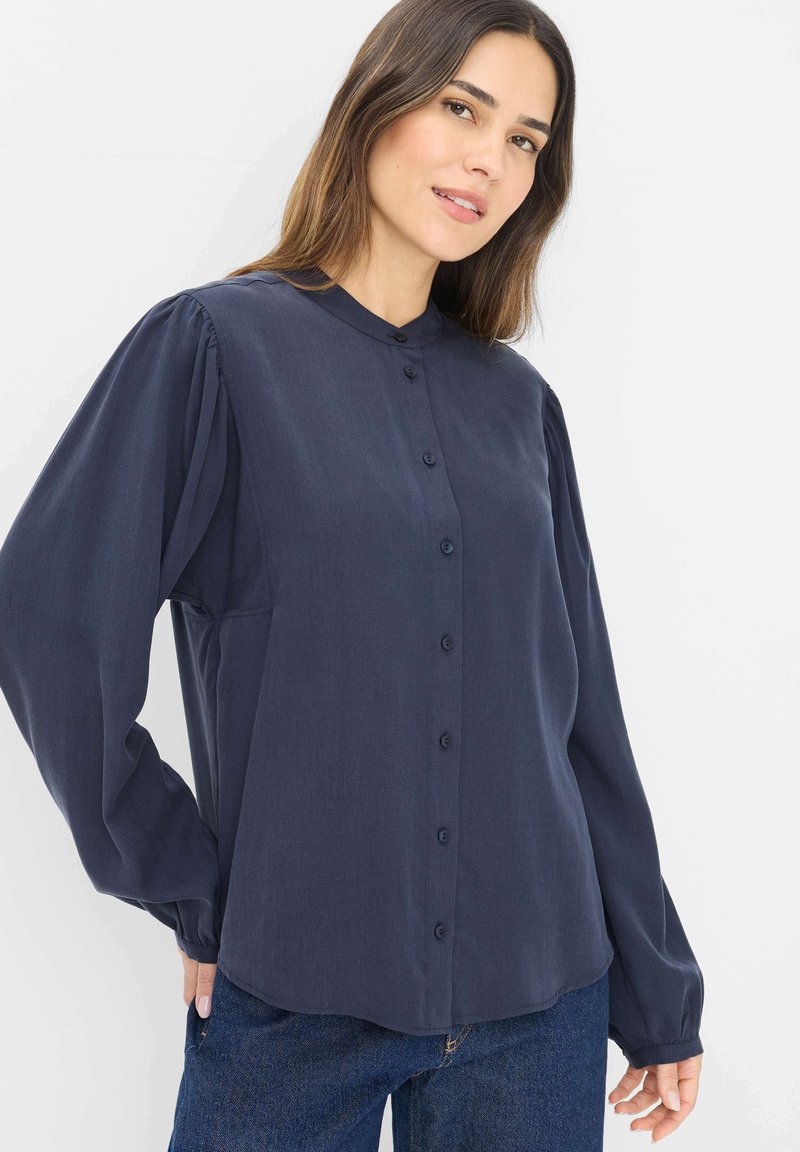 Mørkeblå langærmet bluse med knaplukning foran, blød tekstur, plisserede skuldre og rund krave, parret med blå jeans.