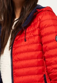 Czerwona puchowa kurtka z granatowym kapturem i zamkami błyskawicznymi. Posiada pikowaną teksturę i sznurek ściągający. Na lewym rękawie znajduje się okrągła naszywka.