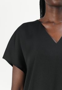 Nő fekete V-nyakú rövid ujjú felsőt visel, fonott haj látható, fehér háttér előtt.