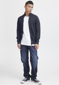Cardigan en maille bleu marine foncé avec un col haut, porté sur un t-shirt blanc uni, associé à un jean en denim bleu et des baskets noires.