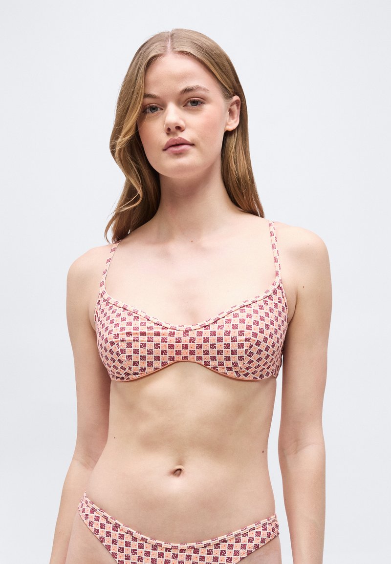 Νεαρή γυναίκα με μακριά μαλλιά, που φοράει μπικίνι με ροζ και κόκκινο σχέδιο, πάνω και κάτω, μπροστά σε απλό ανοιχτό φ όντο.