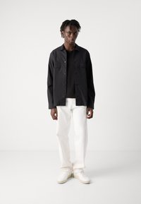 Camisa negra de botones con dos bolsillos en el pecho, combinada con jeans blancos de pierna recta y zapatillas blancas, de pie contra un fondo liso.