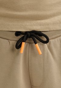 Beige joggebukser med fleksibelt taljebånd, udstyret med en sort snøre med orange ender og sidelommer. Glat stoftekstur.
