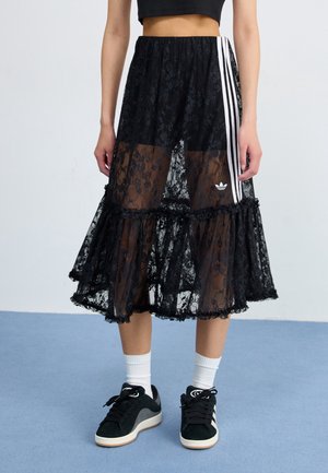 Personne portant une jupe en dentelle noire avec des rayures blanches et un logo, des chaussettes blanches et des baskets noires, debout sur un sol bleu avec un fond clair.