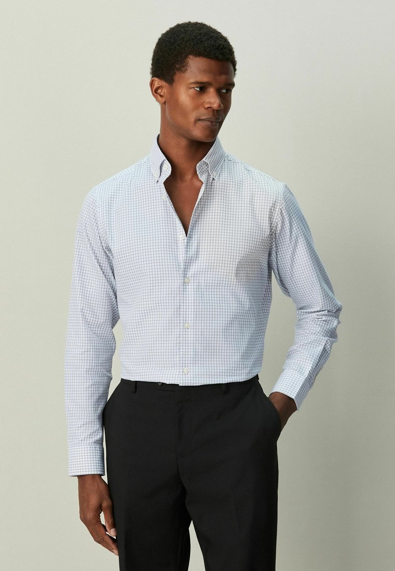 Cămașă cu nasturi în flașă, de culoare albastru deschis, cu model în carouri bleumarin, mâneci lungi și croială ajustată. Asortată cu pantaloni întunecați, stând într-un decor neutru.