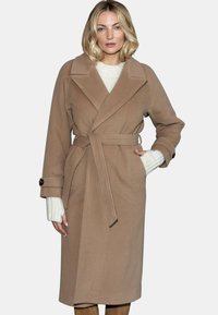 Długi beżowy płaszcz z miękkiego materiału, z szerokimi klapami, przewiązanym wąsem i guzikami na rękawach. Modelka ma na sobie biały sweter.