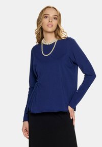 Granatowa bluzka z długim rękawem, okrągłym dekoltem i marszczeniem z boku, w zestawie z czarną spódnicą. Charakteryzuje się gładką, miękką teksturą.