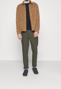 Brązowa kurtka z sztruksu, granatowy sweter, oliwkowe spodnie i ciemnoniebieskie buty. Z elementami takimi jak guziki, kieszenie i podwinięte mankiety spodni.