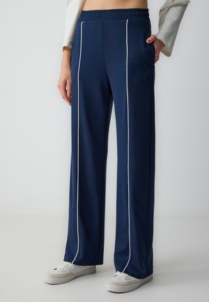 Marineblauwe trainingsbroeken met een relaxte pasvorm, voorzien van witte zijpiping, elastische tailleband, zachte textuur en gestyled met witte sneakers.