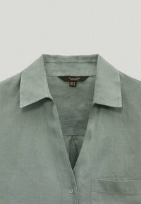 Chemise en lin vert clair avec un col pointu, une fermeture à un bouton et une poche poitrine. Le tissu a une apparence douce et texturée.