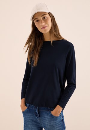 Haut à manches longues bleu marine en tissu doux, associé à un jean bleu clair. Le modèle porte une casquette gris clair, se tenant avec les mains dans les poches.