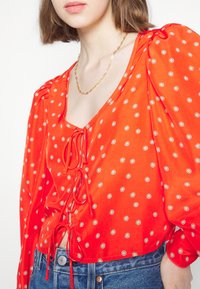 Blusa vermelha com mangas bufantes e detalhe de laço na parte da frente. Apresenta bolinhas brancas e amarelas num tecido suave. Combinada com denim.