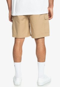 Shorts cargo beiges avec un motif texturé, des poches latérales et une coupe décontractée, portés avec des chaussettes blanches et des baskets.