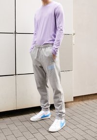 Ljuslila långärmad tröja, grå mjukisbyxor med sidostripes och blått Nike-logotyp, vita sneakers med blå detaljer, stående på asfalt.