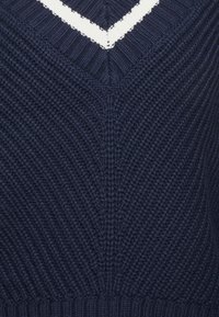 Granatowy sweter z dzianiny z dekoltem w kształcie litery V i białym akcentującym paskiem. Posiada teksturowane ściągacze oraz zstępujący wzór chevron na przodzie.