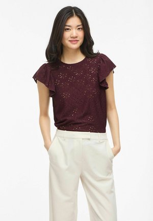 Młoda kobieta o ciemnych włosach, ubrana w bordowy top z ażurowej koronki z falbankowymi rękawami oraz kremowe wysokowalne spodnie, ręce w kieszeniach.