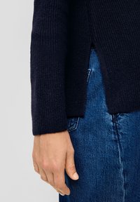 Marine gebreide trui met een gestructureerde stof, voorzien van zijsplitten. Gecombineerd met blauwe denim, met een close-up van de mouw en hand.
