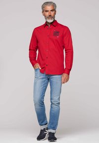 Rotes Hemd mit Knöpfen und blauen Knöpfen sowie dem "CAMP DAVID"-Logo auf der Brust, kombiniert mit hellblauen Jeans und dunklen Sneakers.