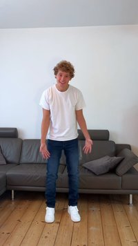 Balta apvalaus kaklo marškinėlius, mėlynus džinsus ir baltus sportbačius. Stovi ant medinės grindų, šalia pilko sofą su dviem raštuotais pagalvėlėmis.