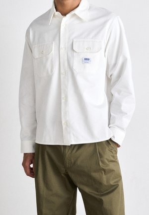 Mężczyzna ubrany w białą koszulę zapinaną na guziki z dwoma kieszeniami na piersi, jedną z małą naszywką z logo Hugo, w połączeniu z oliwkowozielonymi spodniami.