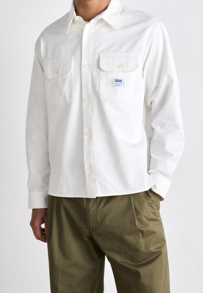 Homme portant une chemise blanche à boutons avec deux poches poitrine, dont l'une comporte un petit écusson du logo Hugo, associée à un pantalon vert olive.