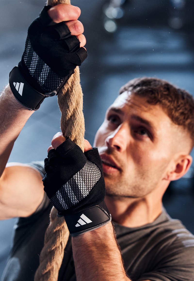 Black training gloves with mesh and grip accents, gripping a rope. Appears in a fitness setting with a dark background and focused expression.