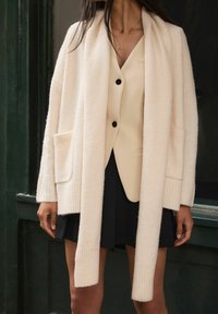 Lichtbeige cardigan met geribbelde manchetten en zakken, gemaakt van zachte stof, gecombineerd met een crèmekleurige blazer en een zwarte rok. Eenvoudig ontwerp.