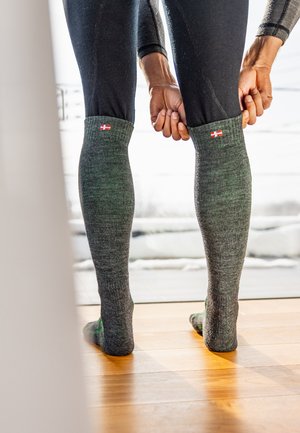 Graue knielange Socken mit geripptem Textur und einem kleinen rot-weißen Etikett, getragen von einer Person in schwarzen Leggings, die auf einem Holzboden steht.