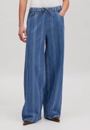 Person trägt weit geschnittene blaue gestreifte Jeans mit elastischem Bund und Kordelzug, kombiniert mit braunen spitzen Schuhen.