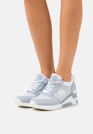 Jambes portant des baskets à lacets bleu clair et blanc avec un logo sur le côté, debout sur une surface blanche plate.