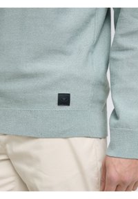 Suéter verde claro hecho de tela texturizada, con puños acanalados y un pequeño parche de logo negro en el costado. Combinado con pantalones de color claro.