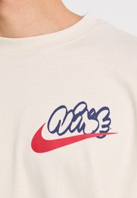 Krémszínű póló, amelyen egy nagy kék "VIBE" grafika és egy piros Nike swoosh logó található az elején. Az anyag simának és könnyűnek tűnik.