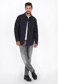 Marineblaues Hemd -Jackett mit Knopfleiste, zwei Brusttaschen und Schulterklappen; kombiniert mit grauen Jeans und schwarzen Knöchelschuhen. Glatte Stoffbeschaffenheit.