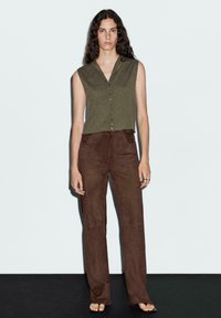 Top senza maniche verde oliva con scollatura a V e chiusura con bottoni, abbinato a pantaloni svasati in suede marrone. Tessuto liscio, dettagli minimi.