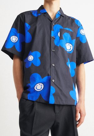 Zwart overhemd met korte mouwen en knopen, met grote blauwe bloemen met witte glimlachende gezichten en de letters H, G en O, gedragen met een zwarte broek.