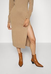Kamel farbenes Strickkleid mit langen Ärmeln und seitlichem Schlitz; kombiniert mit braunen spitz zulaufenden Knöchelstiefeln mit einem seitlichen Reißverschluss.
