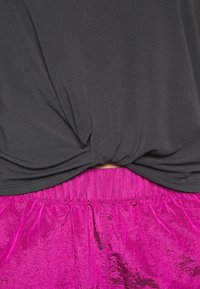 Camiseta negra con un detalle de nudo en la parte delantera sobre pantalones cortos plisados de un vibrante color rosa, hechos de un tejido brillante y con una cintura elástica.