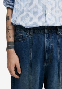 Blau-weiß gestreiftes Hemd mit abstraktem Muster, getragen über dunklen Jeans mit Kontrastnähten und Fünf-Taschen-Design.